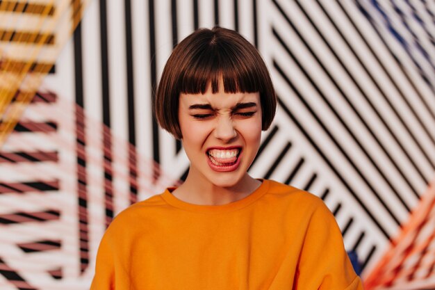 Menina emocional com cabelo castanho e moletom laranja fazendo careta lá fora