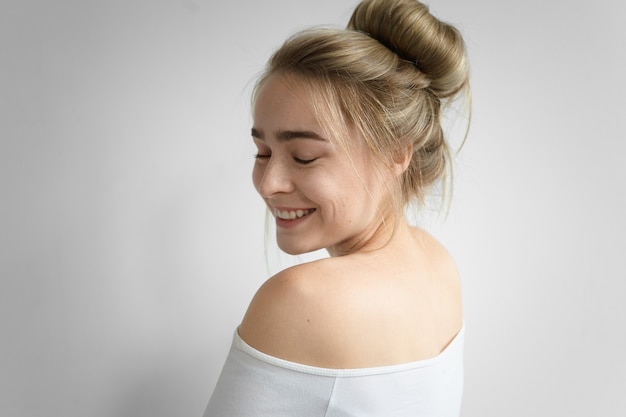 Menina encantadora com coque de cabelo e costas nuas posando isoladas, fechando os olhos e sorrindo amplamente. foto de uma atraente jovem caucasiana com penteado bagunçado, aproveitando um bom dia