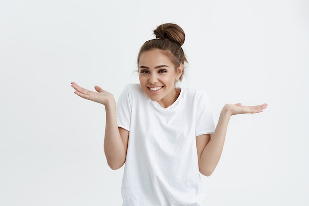 Menina encantadora com coque encolher os ombros, espalhando as mãos em gesto sem noção, sorrindo amplamente, estar de bom humor