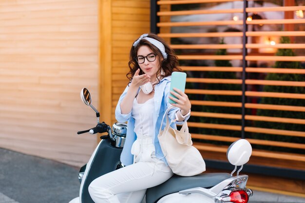 Menina encantadora com penteado encaracolado mandando beijo no ar para a foto, enquanto está sentada na scooter na rua