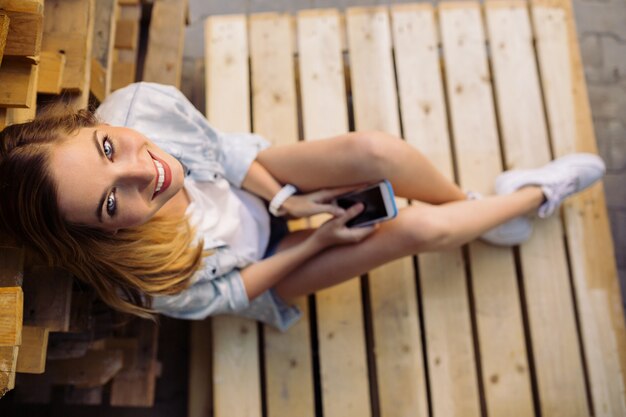 Menina encantadora, sentado em um palete usando telefone inteligente