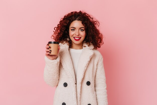 Menina encaracolada de olhos azuis com batom vermelho, vestida com casaco de pele branca eco, sorrindo e segurando um copo de café no espaço rosa.