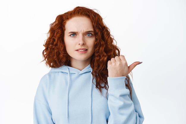 Menina encaracolada ruiva confusa mostrando algo estranho apontando para a direita e franzindo a testa duvidosa em pé hesitante e intrigada contra o fundo branco