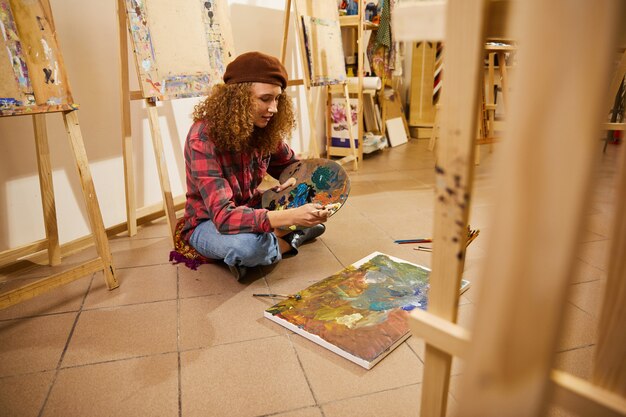 Menina encaracolada senta-se no chão e desenha uma pintura com óleos