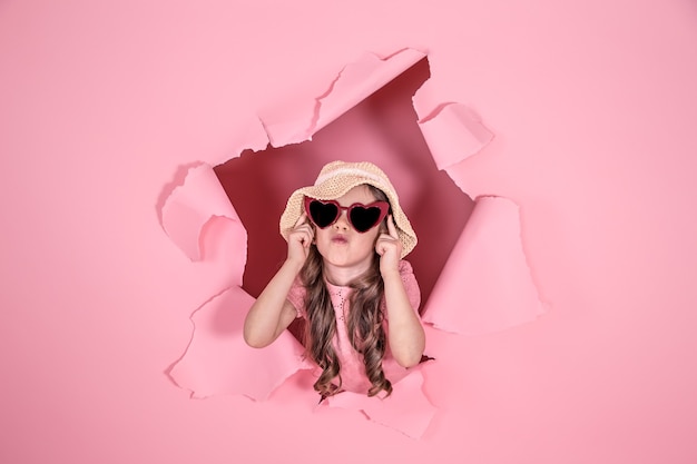 Foto grátis menina engraçada espiando de um buraco com um chapéu de praia e óculos em forma de coração em um fundo colorido, lugar para texto, estúdio de tiro