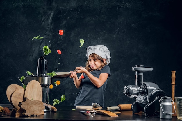 Menina engraçada está jogando legumes na panela no estúdio fotográfico escuro.