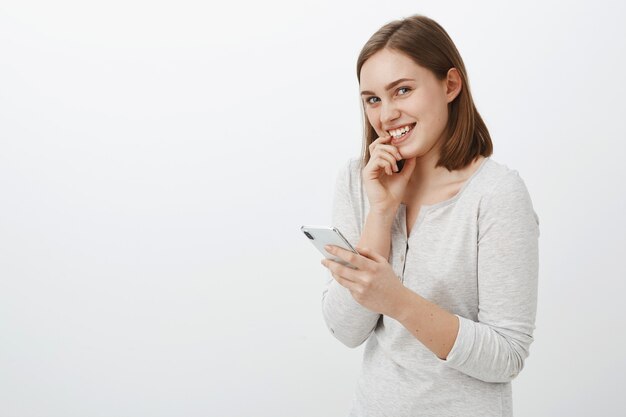 Menina enviando mensagem arriscada para cara legal rindo glamour e olhando sorrindo alegremente, segurando o dedo tímido no lábio e o smartphone na mão, parada divertida e animada sobre a parede cinza