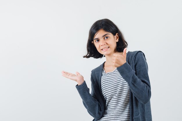 Menina espalhando a palma da mão para o lado, aparecendo o polegar em uma camiseta, jaqueta e parecendo confiante.