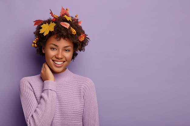 Menina étnica encantada expressa emoções sinceras, toca o pescoço e ri alegremente, vestida com agasalho de malha quente, olha com largo sorriso, tem penteado com folhas amarelas