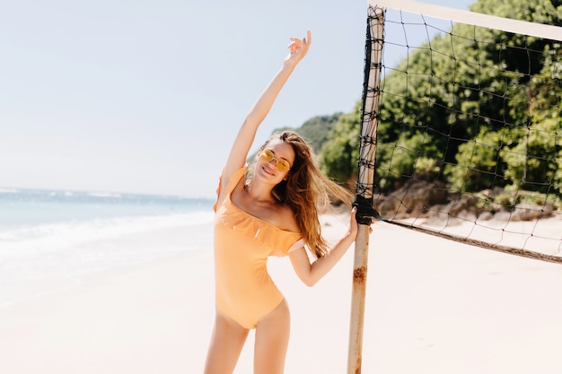 Menina europeia bem torneada com cabelo castanho dançando na ilha tropical em férias. Foto ao ar livre do feliz modelo feminino em trajes de banho tocando o conjunto de vôlei.