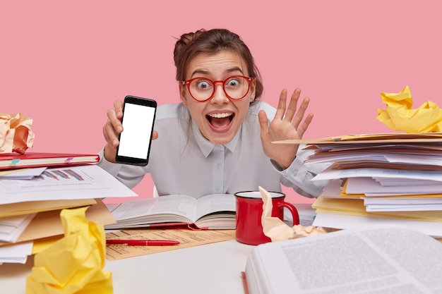 Foto grátis menina europeia sorridente e encantada acenando com a mão, segurando o celular com tela de simulação, animada, bebe bebida quente