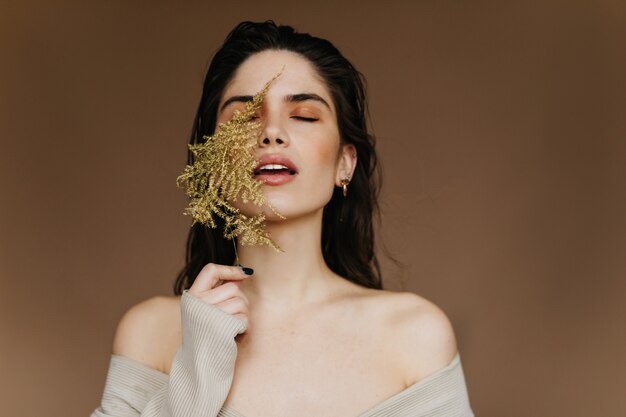 Menina fascinante segurando folha verde e em pé com os olhos fechados. Mulher relaxada elegante que aprecia a sessão de fotos interna.