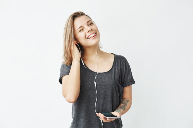 Menina feliz bonita nova que sorri piscando mostrando a música de escuta da língua nos fones de ouvido.