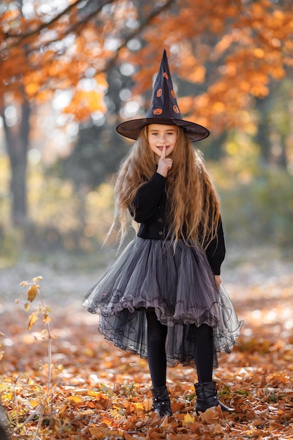 Menina feliz bonitinha vestida com fantasia de bruxa em pé com fundo de floresta de outono. menina usando vestido e chapéu de cone. garota vestida para o halloween.