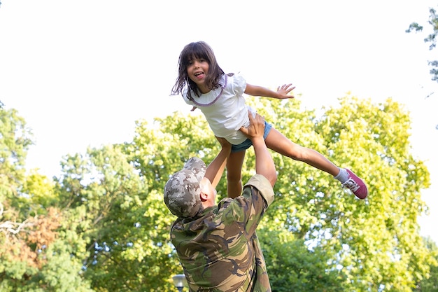 Menina feliz brincando com o pai em uniforme militar. Vista traseira do pai levantando a filha.