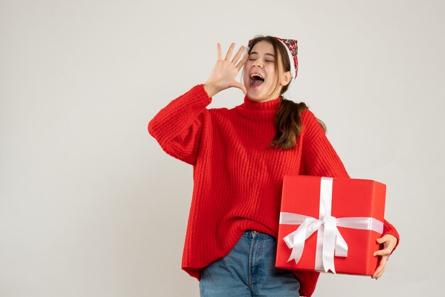 menina feliz com chapéu de Papai Noel segurando um presente gritando em pé no branco