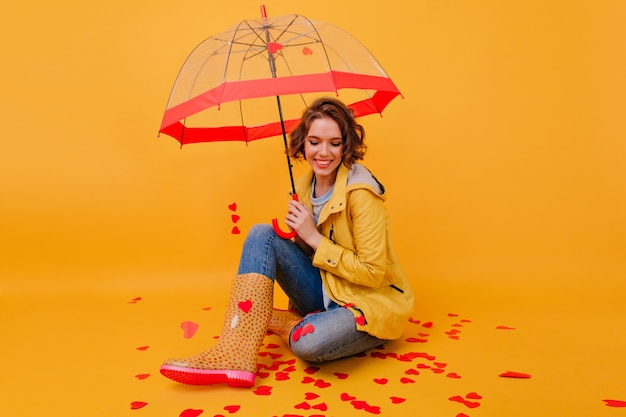 Menina feliz com sapatos de borracha, sentada com guarda-chuva no chão e rindo. feliz mulher branca com casaco de outono, aproveitando o dia dos namorados.