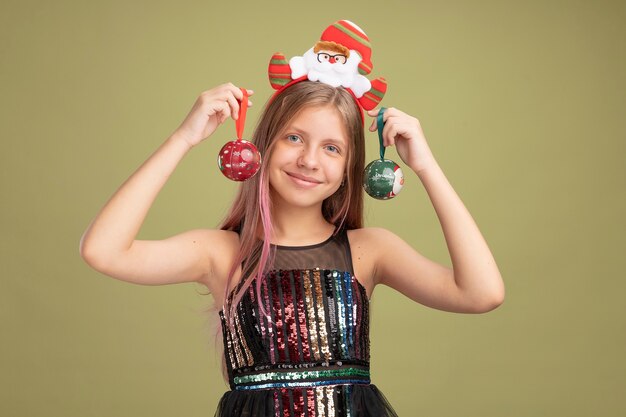 Menina feliz com vestido de festa glitter e bandana com o Papai Noel segurando bolas de Natal, olhando para a câmera com um sorriso no rosto em pé sobre um fundo verde