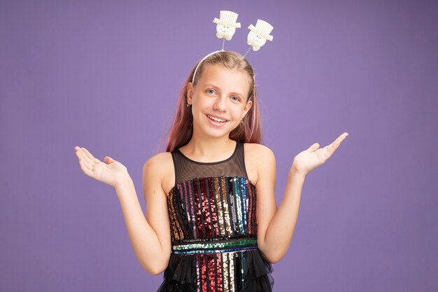 Menina feliz com vestido de festa glitter e bandana engraçada olhando para a câmera sorrindo com os braços levantados em pé sobre um fundo roxo