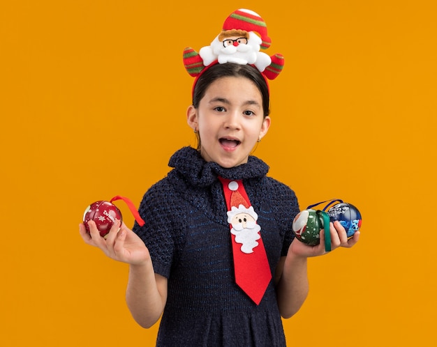 Menina feliz e animada em um vestido de malha com gravata vermelha com aro engraçado na cabeça segurando bolas de natal e sorrindo alegremente