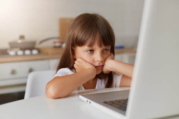 Menina feliz e fofa de 8 anos de idade em uma camiseta listrada e jeans com óculos, sentada em um tapete na frente de um laptop, tecnologias de educação remota e dever de casa