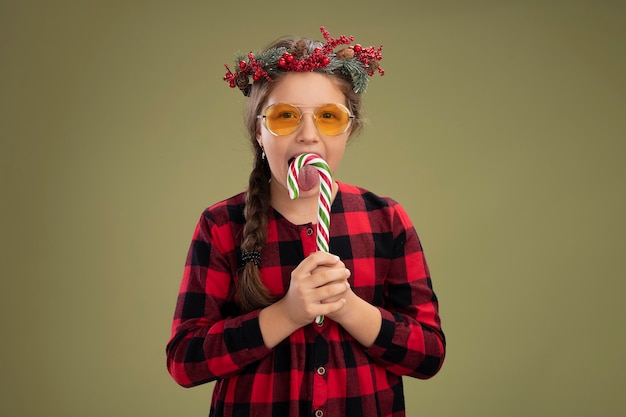 Menina feliz e positiva usando guirlanda de natal em vestido xadrez segurando o bastão de doces, olhando para a câmera sorrindo alegremente indo morder de pé sobre fundo verde