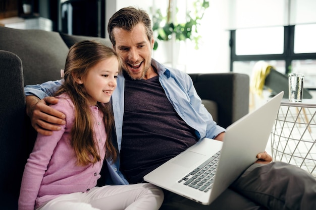 Foto grátis menina feliz e seu pai usando um computador e assistindo algo na internet enquanto relaxa na sala de estar