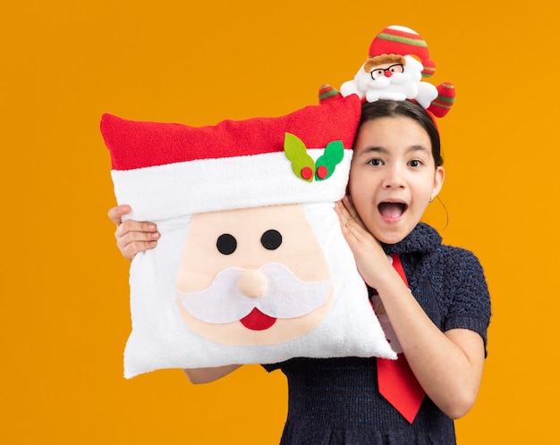 Menina feliz e surpresa com vestido de malha usando gravata vermelha com aro engraçado na cabeça segurando a almofada de natal e sorrindo alegremente