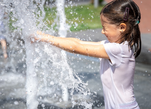 Menina feliz entre os salpicos de água da fonte da cidade se diverte e foge do calor.