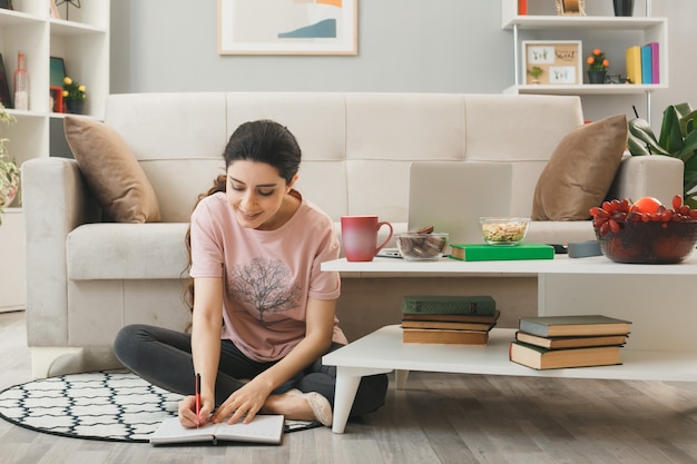 Menina feliz escrevendo no caderno sentado no chão atrás da mesa de centro da sala de estar