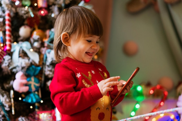 Menina feliz no suéter vermelho segurando o telefone com alegria ao lado da árvore de natal decorada no natal.