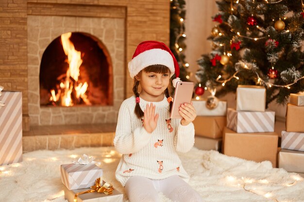 Menina feliz vestindo suéter branco e chapéu de Papai Noel, posando na sala festiva com lareira e árvore de Natal, acenando com a mão para a câmera do celular, tendo a chamada de vídeo.