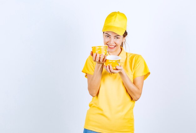 Menina feminina em uniforme amarelo segurando duas xícaras de macarrão para viagem.