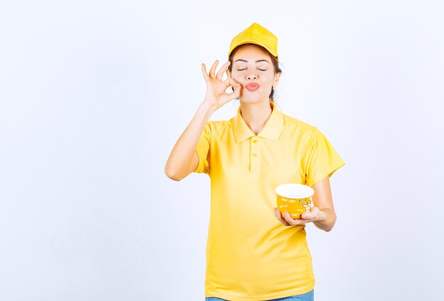 Menina feminina em uniforme amarelo segurando um copo de macarrão amarelo para viagem e mostrando sinal de mão de prazer.