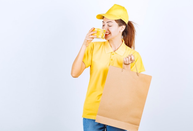 Menina feminina em uniforme amarelo, segurando uma sacola de compras e bebendo uma sopa de macarrão para viagem.