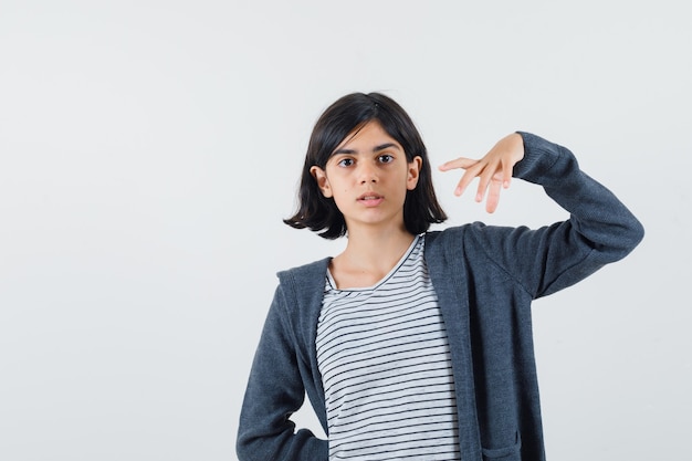 Menina fingindo segurar ou mostrar algo em uma camiseta, jaqueta e parecendo confusa