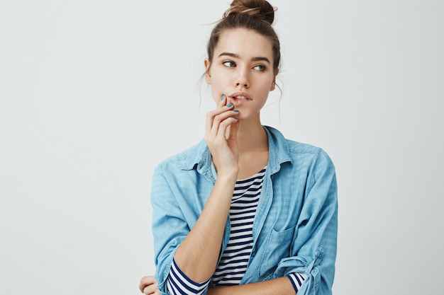 Menina glamourosa elegante com penteado coque, olhando de lado enquanto mordendo o lábio e tocando o queixo, concentrando-se em alguém enquanto sonha ou espaçando, em pé.