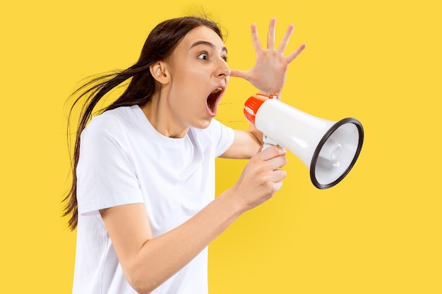 Menina gritando com o porta-voz. Belo retrato feminino de meio comprimento isolado na parede amarela. Jovem mulher sorridente. Espaço negativo. Expressão facial, conceito de emoções humanas.