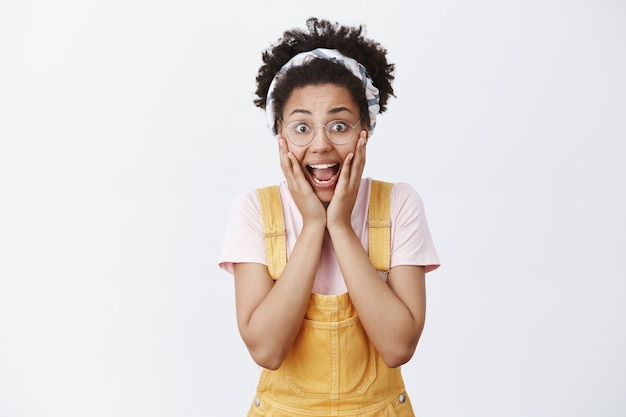 Menina gritando de surpresa. mulher afro-americana fofa e animada espantada de óculos, faixa na cabeça e macacão amarelo, gritando com o queixo caído