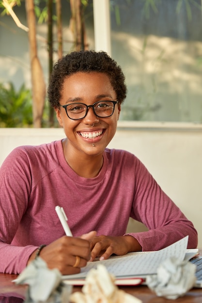 Foto grátis menina hipster positiva com cabelo curto encaracolado, sorri amplamente, usa piercing, escreve a ideia criativa no bloco de notas, envolvida no processo de trabalho, rodeada de bolas de papel, senta-se no local de trabalho sozinha