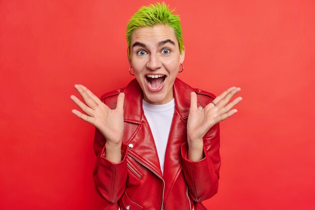 Menina hipster positiva com cabelo verde curto espalha as palmas das mãos exclama de felicidade reage a notícias impressionantes não posso acreditar em revelação chocante usa jaqueta de couro poses contra parede vermelha
