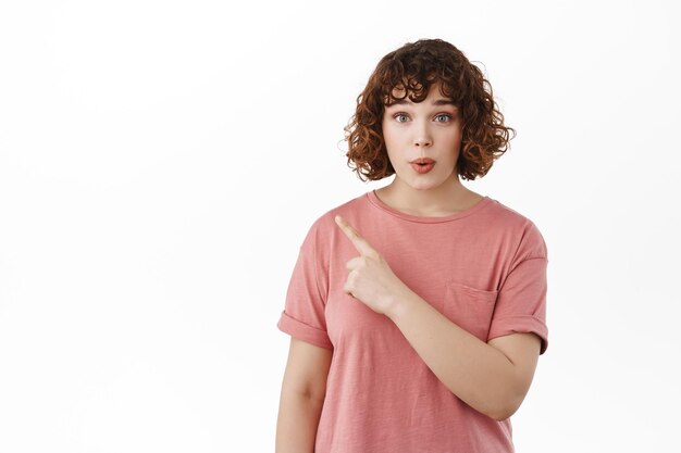 Menina impressionada apontando o dedo no canto superior esquerdo, mostrando propaganda, sendo curiosa e fazendo perguntas sobre acordo promocional, de pé na camiseta contra fundo branco