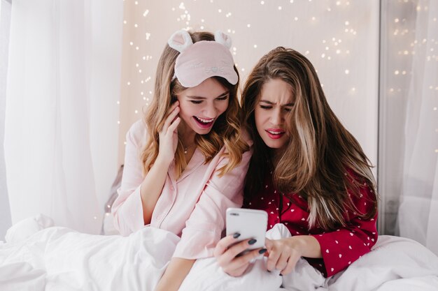 Menina infeliz com penteado comprido, lendo mensagem de telefone na cama. Duas amigas brancas relaxando no quarto com luz interior.