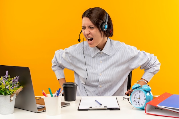 Menina irritada do call center usando fone de ouvido, sentada na mesa com os olhos fechados, isolado em laranja