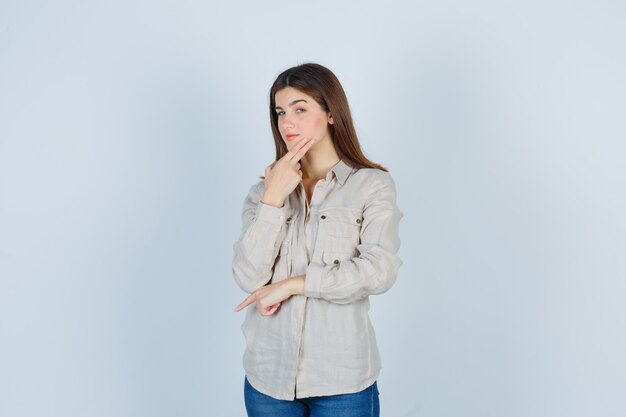 Menina jovem apoiando o queixo na mão, pensando em algo, apontando para baixo com o dedo indicador em uma camisa bege, jeans e olhando pensativa, vista frontal.