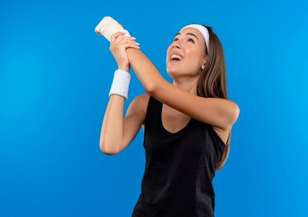 Menina jovem e bem desportiva dolorida usando bandana e pulseira, levantando e segurando o pulso ferido enrolado em uma bandagem e olhando para cima, isolada na parede azul