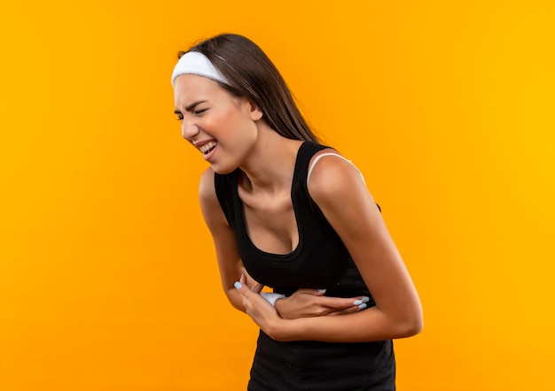 Menina jovem e bem desportiva dolorida usando bandana e pulseira segurando a barriga, sofrendo de dor com os olhos fechados, isolada na parede laranja