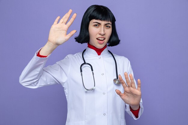 Menina jovem e bonita caucasiana irritada com uniforme de médico e estetoscópio em pé com as mãos levantadas