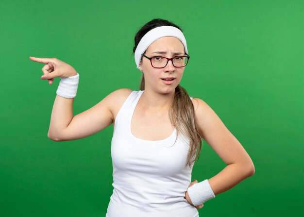 Menina jovem e desportiva insatisfeita com óculos ópticos, fita para a cabeça e pulseiras apontando para o lado