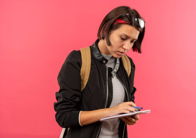 Menina jovem estudante concentrada usando óculos na cabeça e na bolsa, segurando uma caneta e um bloco de notas, olhando para um bloco de notas isolado em rosa com espaço de cópia